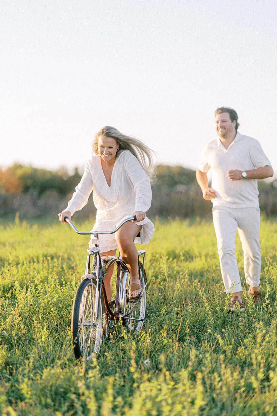 A joyful woman riding a bicycle in a sunlit field with a man running behind her.