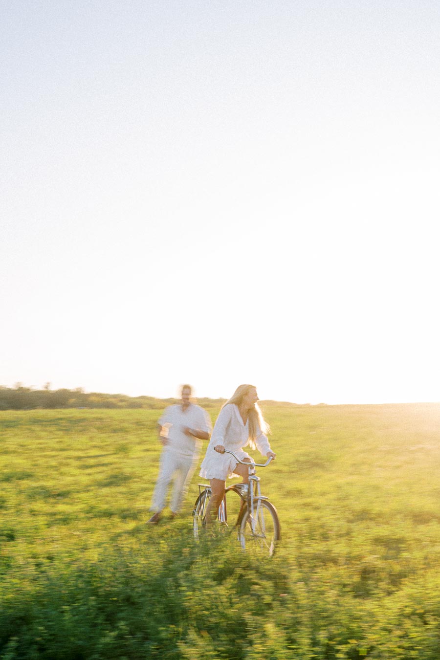 A woman riding a bicycle through a sunny, green field with a man jogging behind her, both enjoying an outdoor activity under a clear, bright sky.
