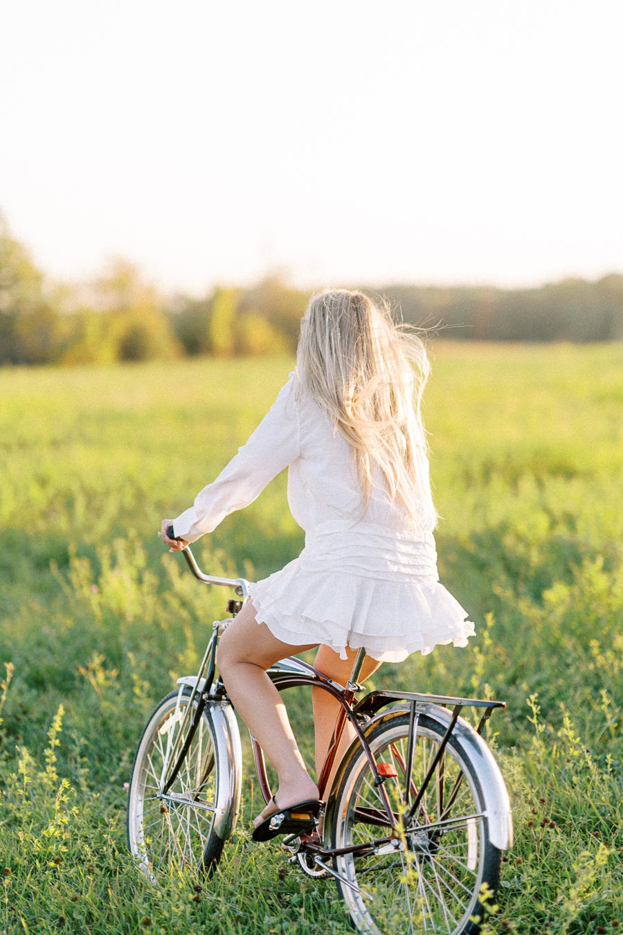 A woman with long blonde hair rides a vintage bicycle through a sunlit green meadow, wearing a white dress, capturing the essence of freedom and tranquility in a rural setting.