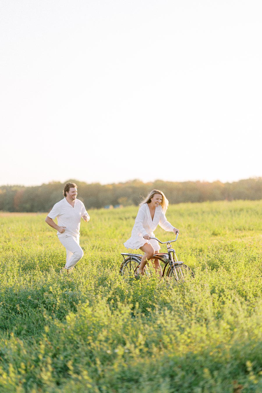 A couple enjoying a sunny day in a lush green field, with a woman riding a bicycle and a man playfully running alongside, both dressed in white.