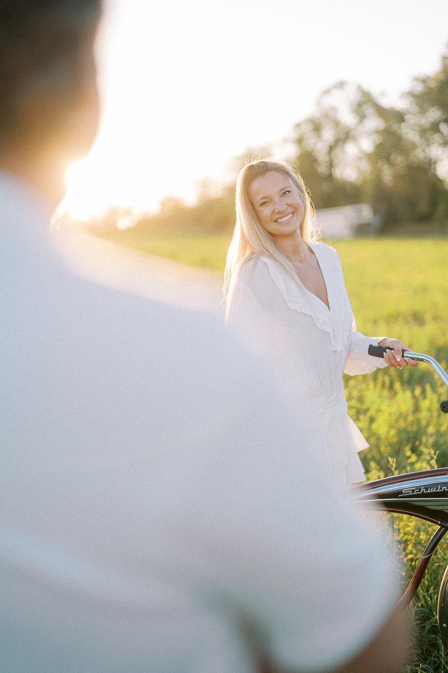 A woman smiling while holding a bicycle in a sunny field, wearing a white outfit during a bright afternoon.
