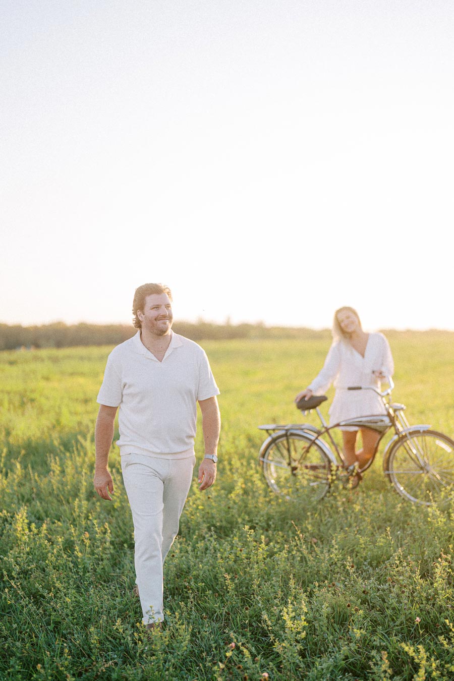 A cheerful man in a white outfit walking through a sunny field, with a woman standing next to a bicycle in the background, surrounded by lush green grass.
