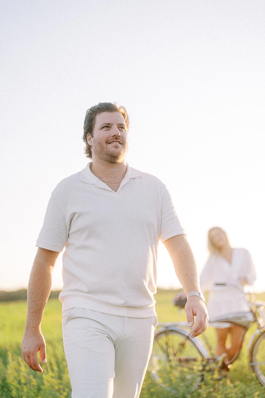 Man in a white outfit walking through a sunlit field, with a woman standing near a bicycle in the background.