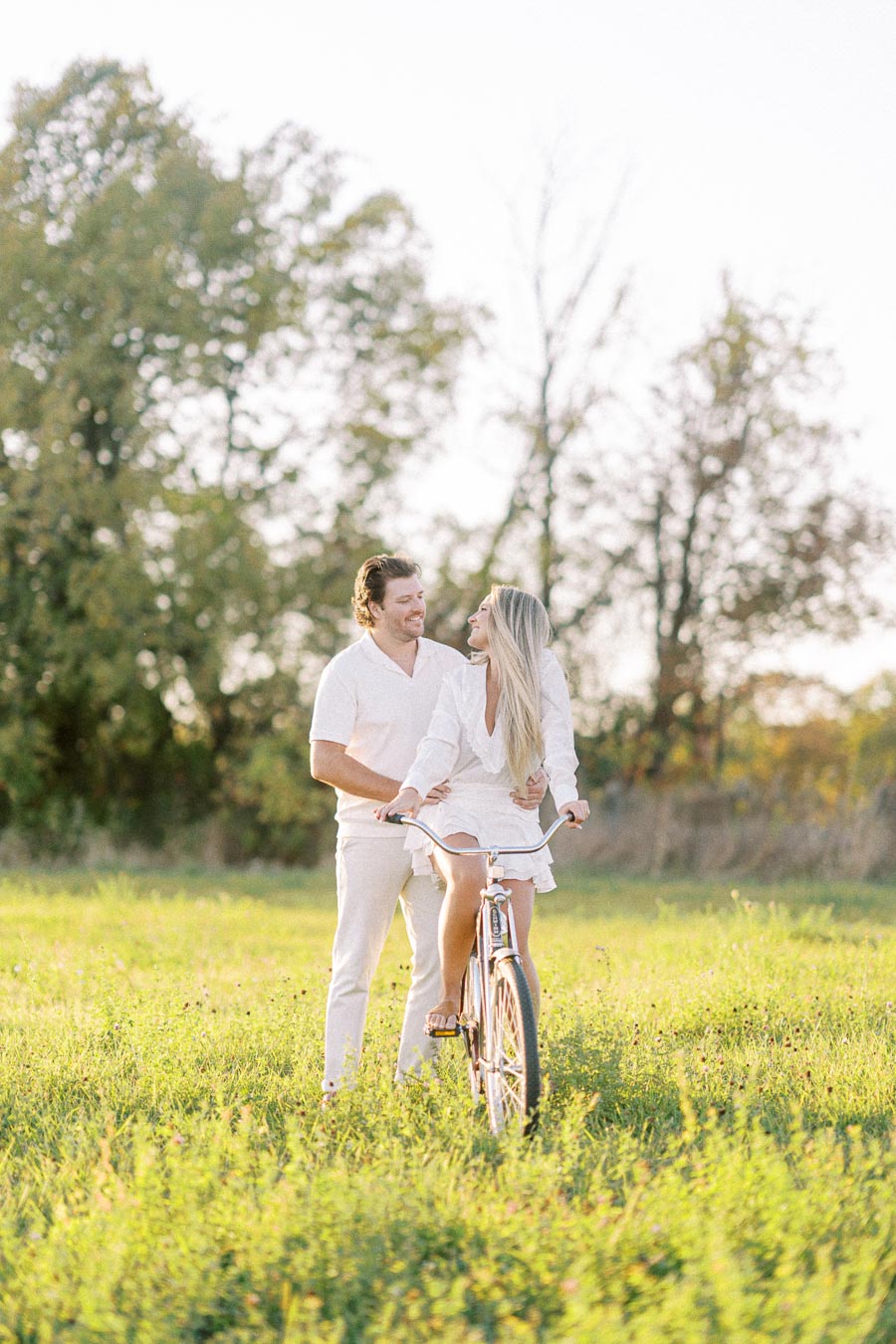 Couple enjoying a sunny day outdoors on a vintage bicycle in a green field with trees in the background.