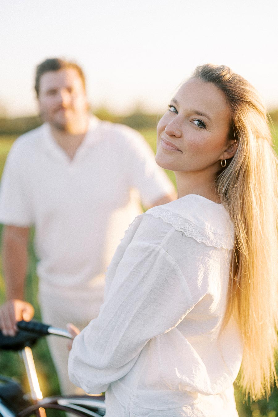 Smiling woman with long blonde hair and white blouse standing outdoors, with a blurred man in the background holding a bicycle, in a sunlit field setting.