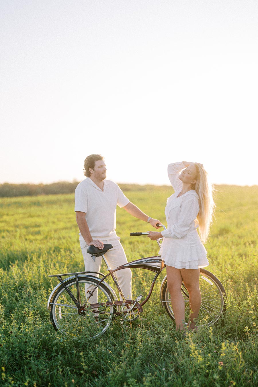 A couple enjoying a sunny day in a lush green field with a vintage bicycle, both dressed in white outfits, smiling and looking at each other, capturing a serene and romantic moment in nature.