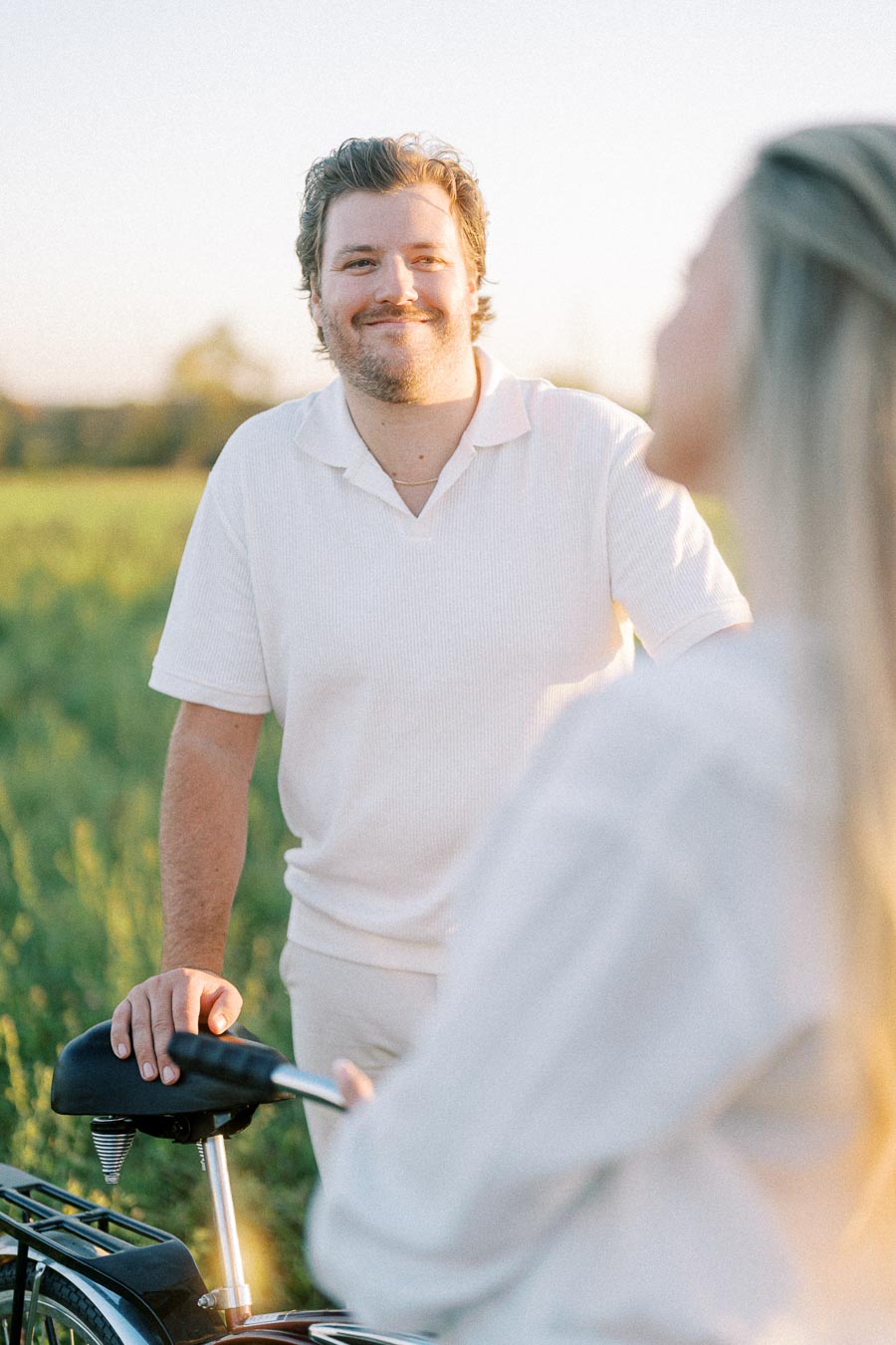 Man smiling in a sunny field next to a bicycle, wearing a white shirt, while engaging in conversation with a blurred woman in the foreground.