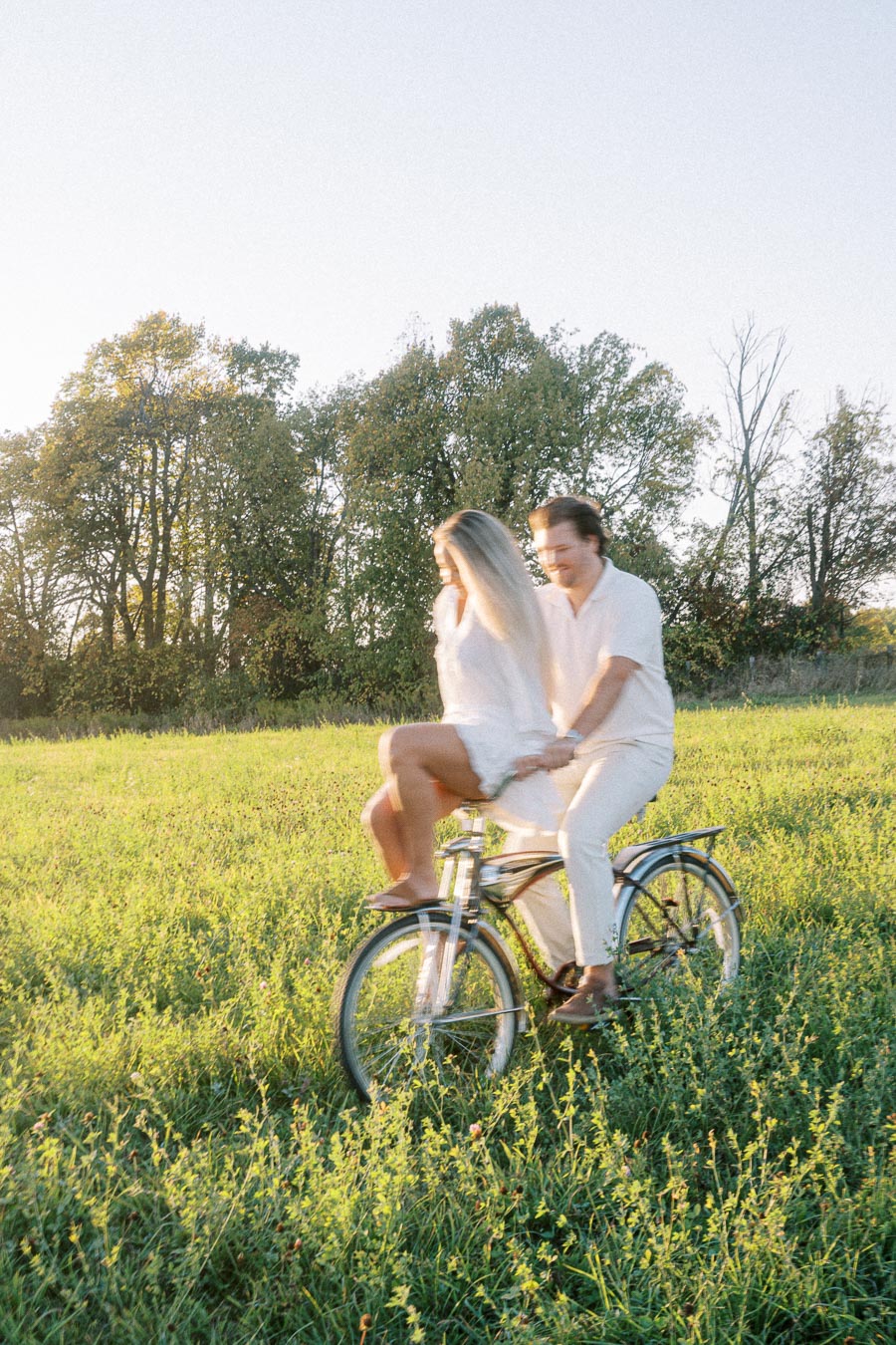 A couple enjoying a tandem bike ride through a lush green field, with bright sunlight filtering through the trees, creating a warm, romantic atmosphere.