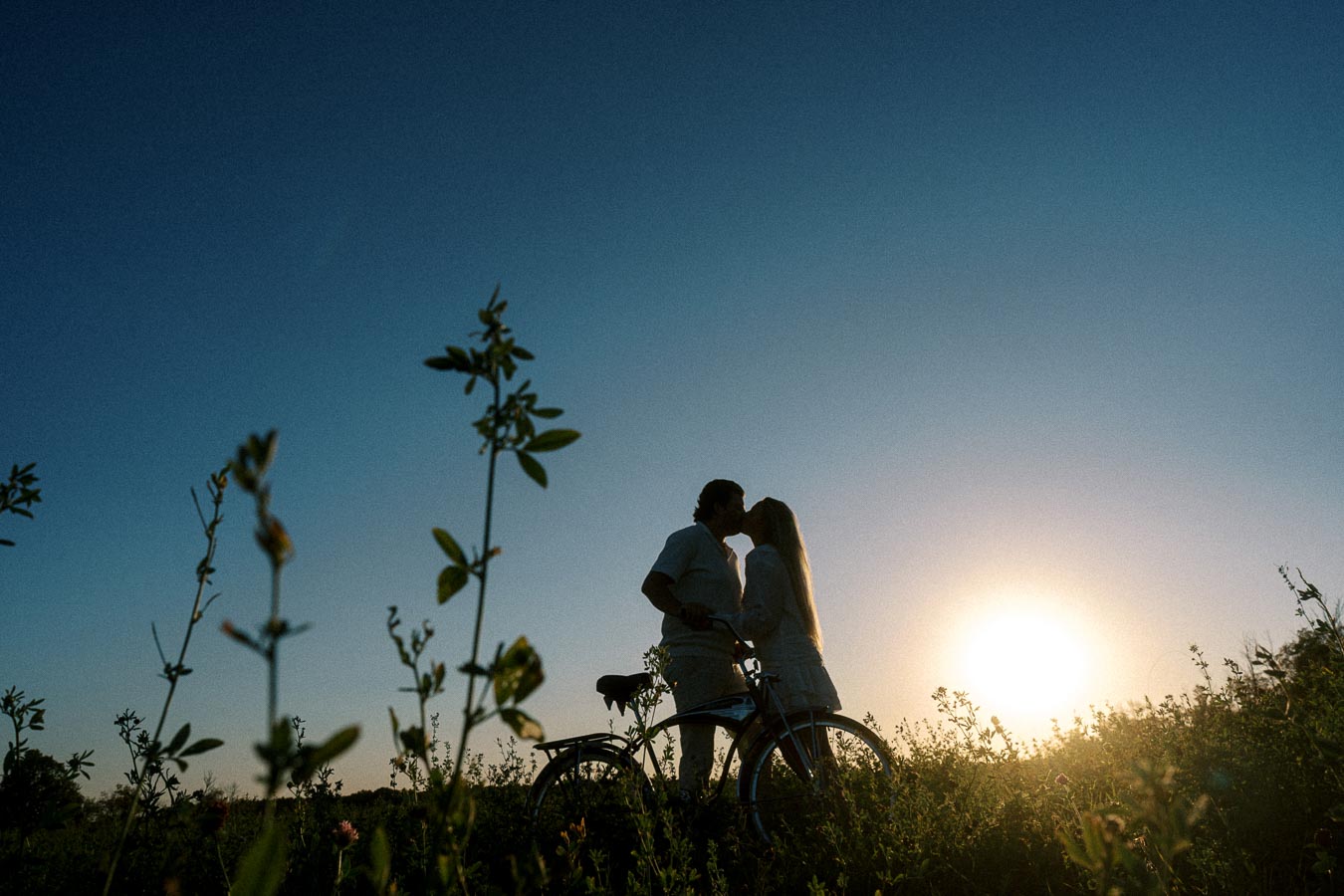 Couple kissing at sunset in a field with a bicycle, surrounded by tall grass and clear blue sky, illustrating a romantic outdoor moment.