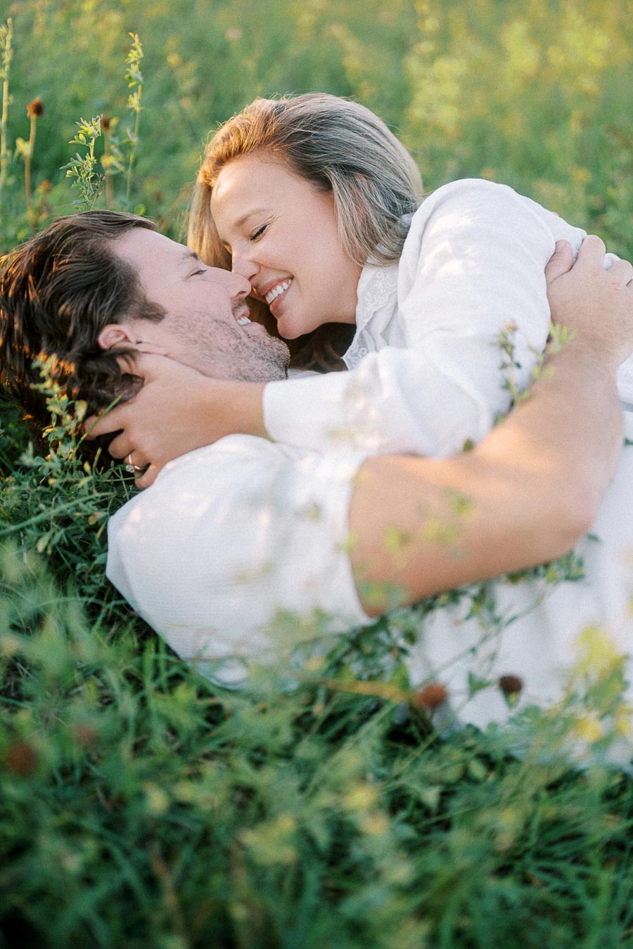 A couple in casual white attire smiling and embracing each other while lying in a lush green meadow, capturing a moment of love and happiness.