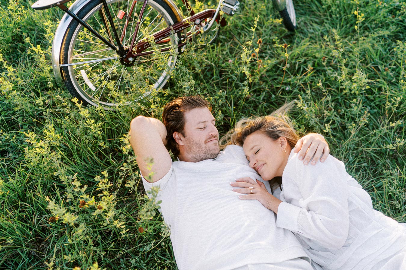 Couple lying on grass next to a bicycle, enjoying a peaceful moment in nature, with the woman resting her head on the man's chest.