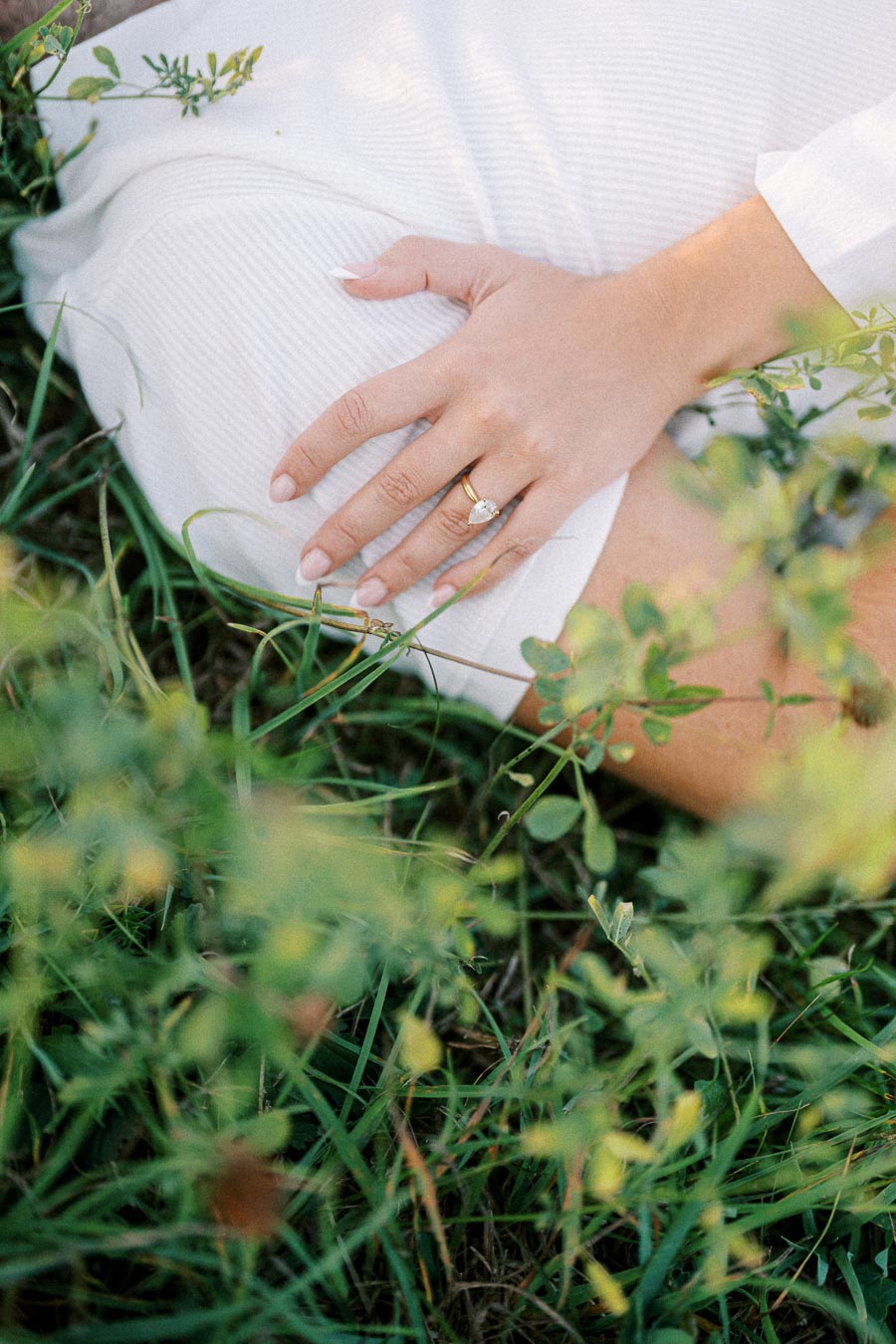 Close-up of a woman's hand resting on her leg surrounded by grass and greenery, featuring a gold ring with a gemstone, conveying a serene and natural setting.