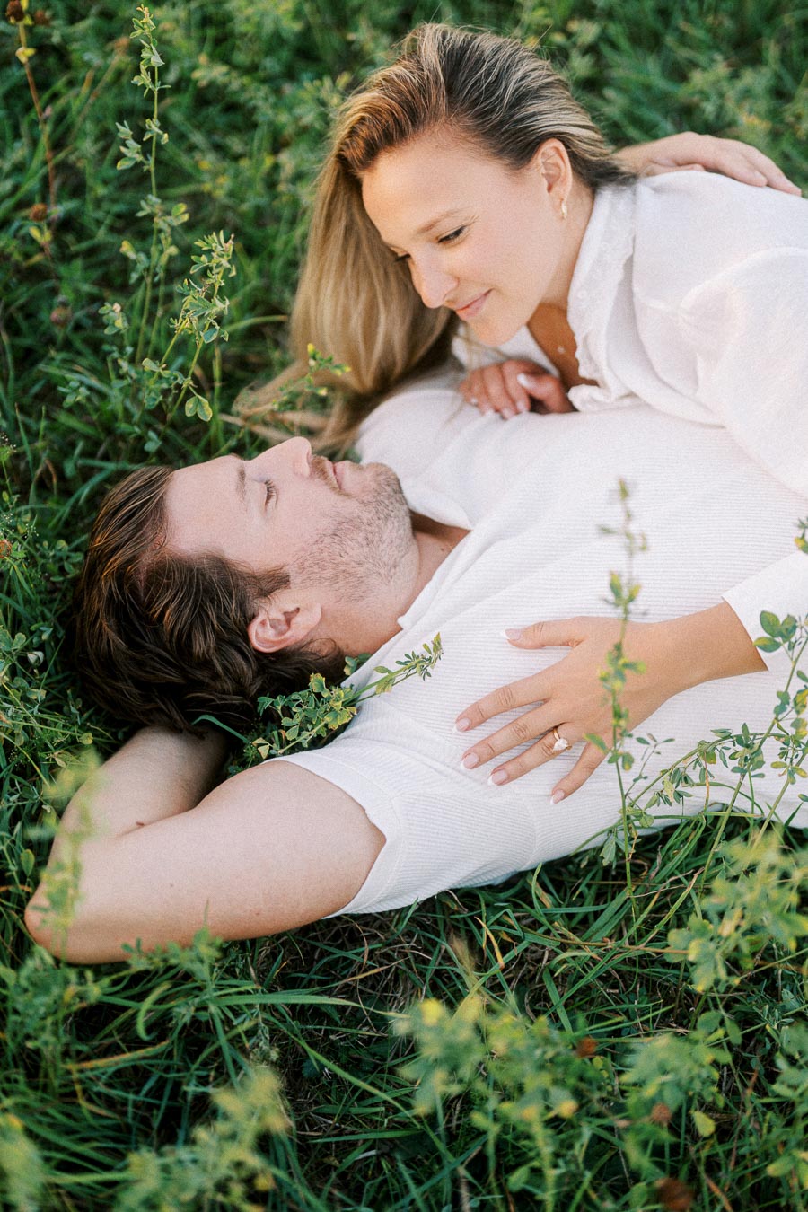 A couple lying in a grassy field, gazing at each other lovingly, wearing casual white clothing.