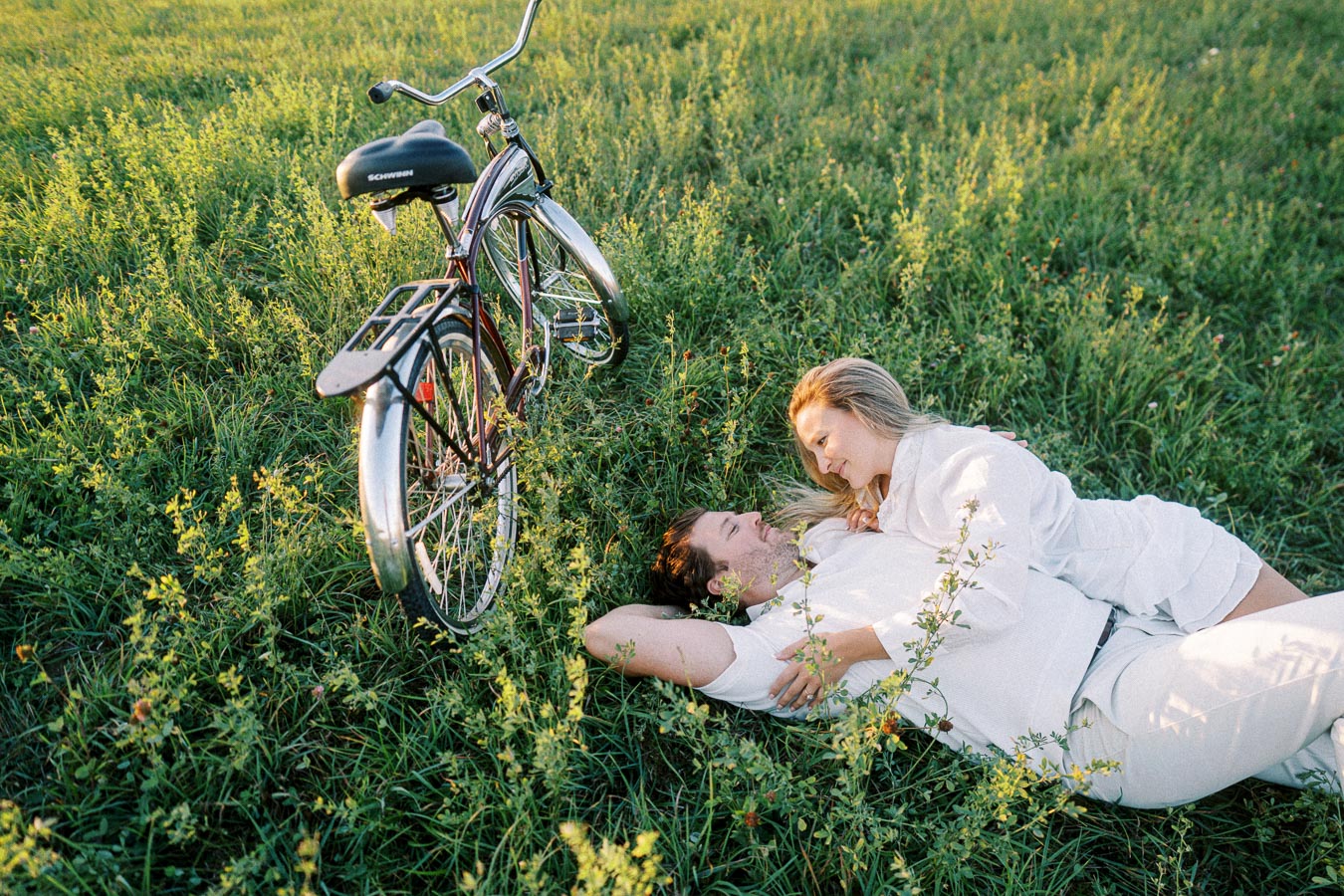 A couple in white clothing enjoying a romantic moment lying in a green field next to a vintage bicycle under the warm sunlight.