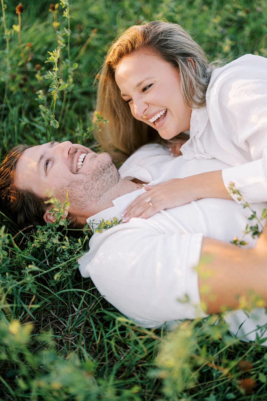 A happy couple lying on grass, sharing a joyful moment together while embracing and smiling, surrounded by greenery and soft sunlight.