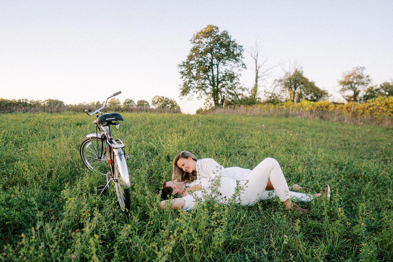 A couple in matching white outfits lies in a lush green field beside a vintage bicycle, surrounded by trees under a clear blue sky, enjoying a peaceful outdoor moment.