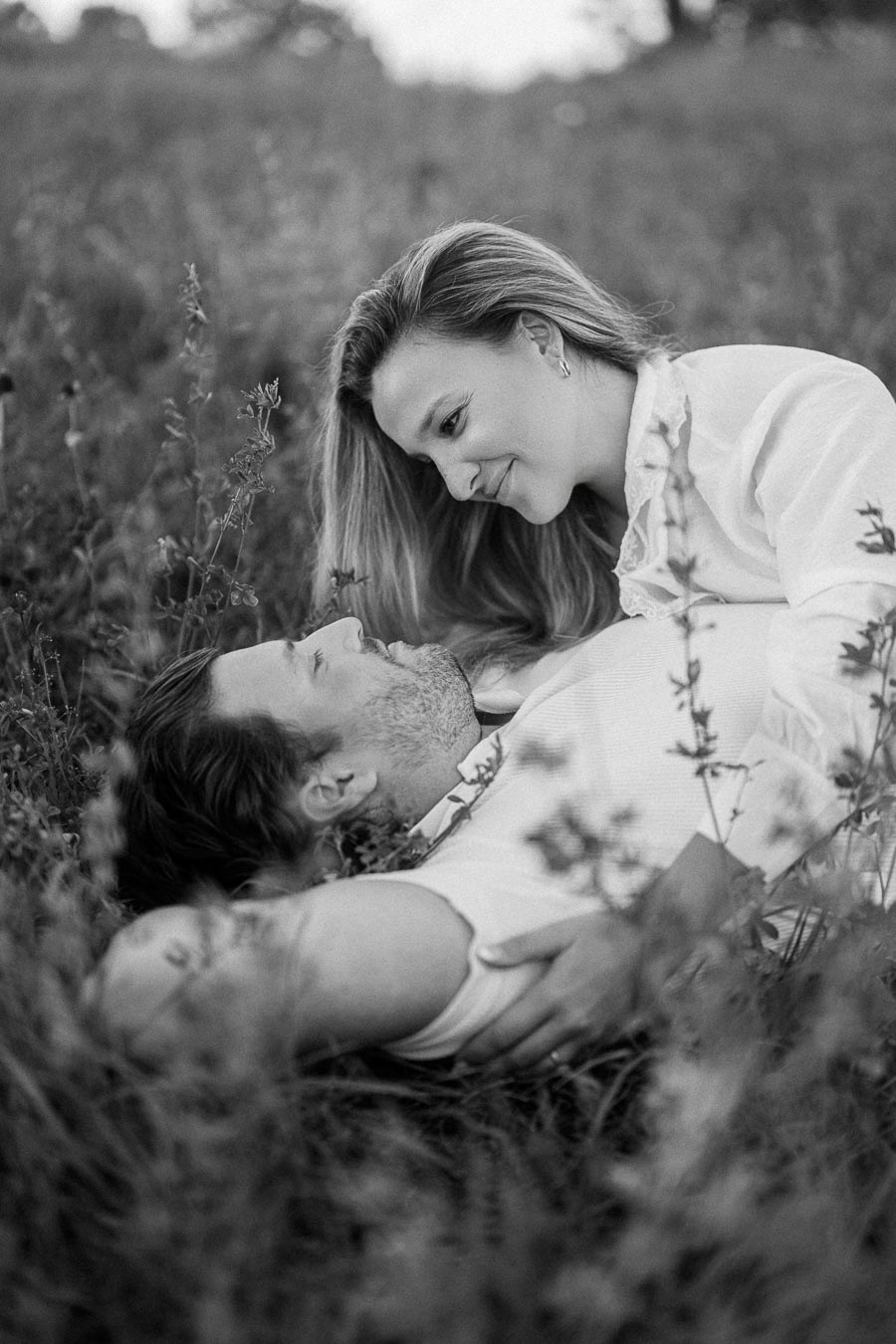 A black and white photo of a couple lying in a grassy field, gazing at each other lovingly, capturing an intimate and romantic moment.