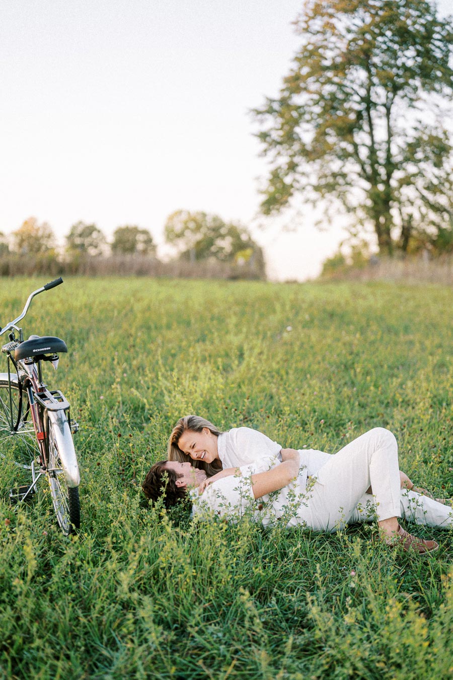 Couple enjoying a romantic moment lying in a grassy field with a bicycle nearby, under a clear sky, surrounded by lush greenery.