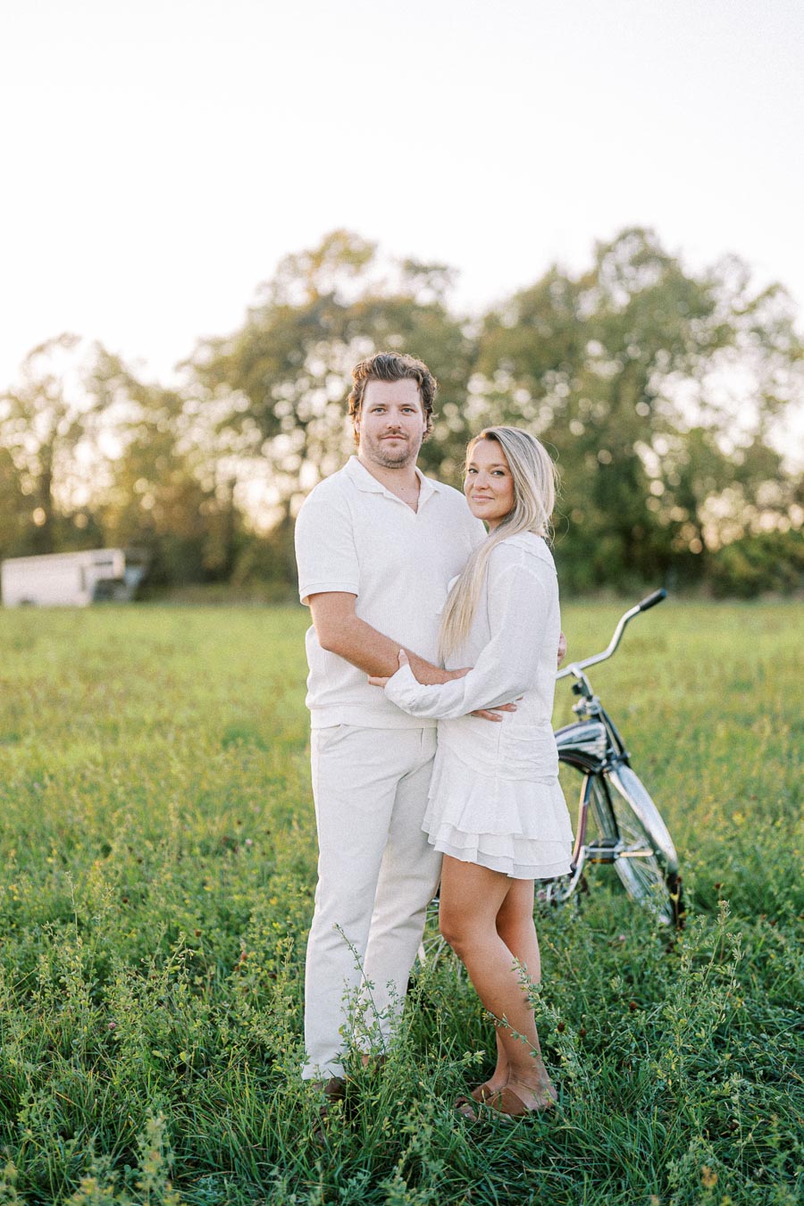 Couple in white outfits embracing in a sunny green field with a vintage bicycle behind them, tall trees in the background