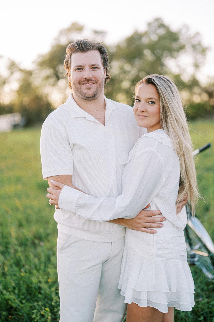 A couple in white clothing embracing in a grassy field with trees in the background during sunset.