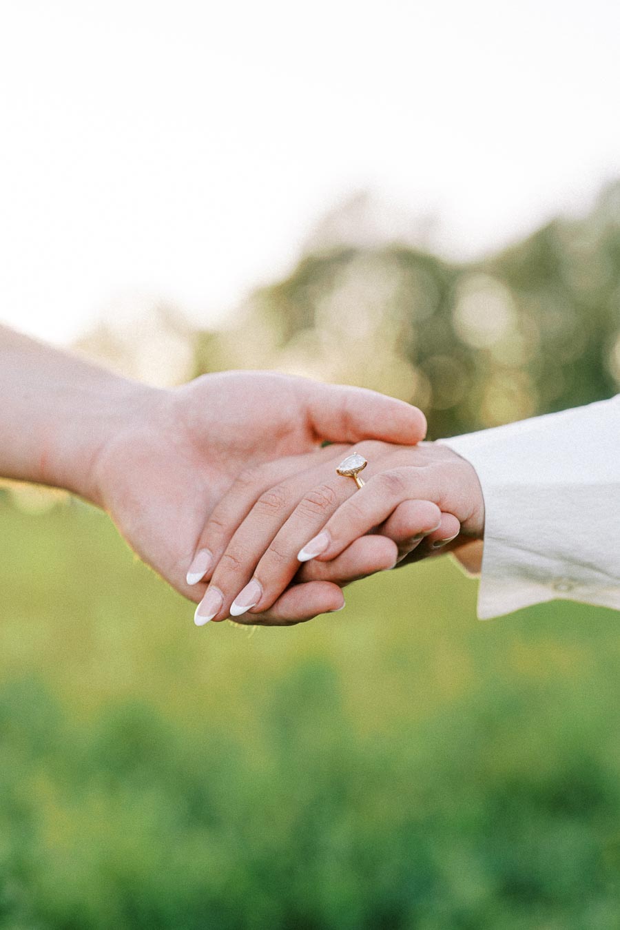 A close-up of two hands gently holding each other, featuring an engagement ring with a large oval stone, set against a blurred outdoor background, symbolizing love and commitment.