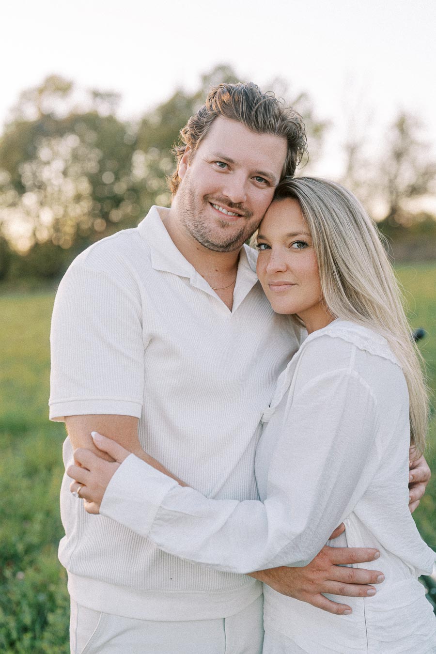 A happy couple embracing in a serene outdoor setting, both wearing white outfits, with lush greenery and trees in the background.