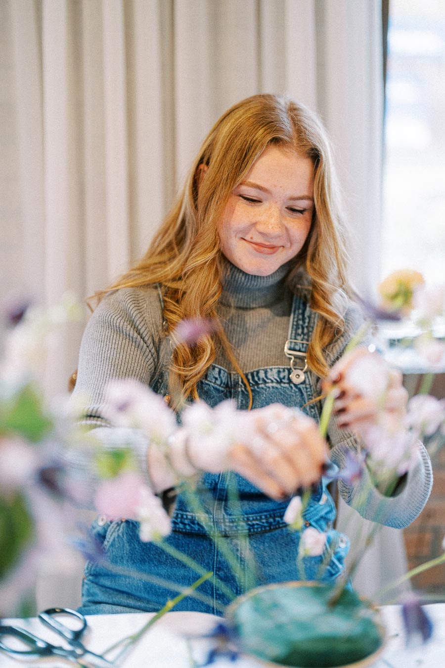 Young woman arranging flowers while smiling, wearing a gray turtleneck sweater and denim overalls, in a bright and cozy room.