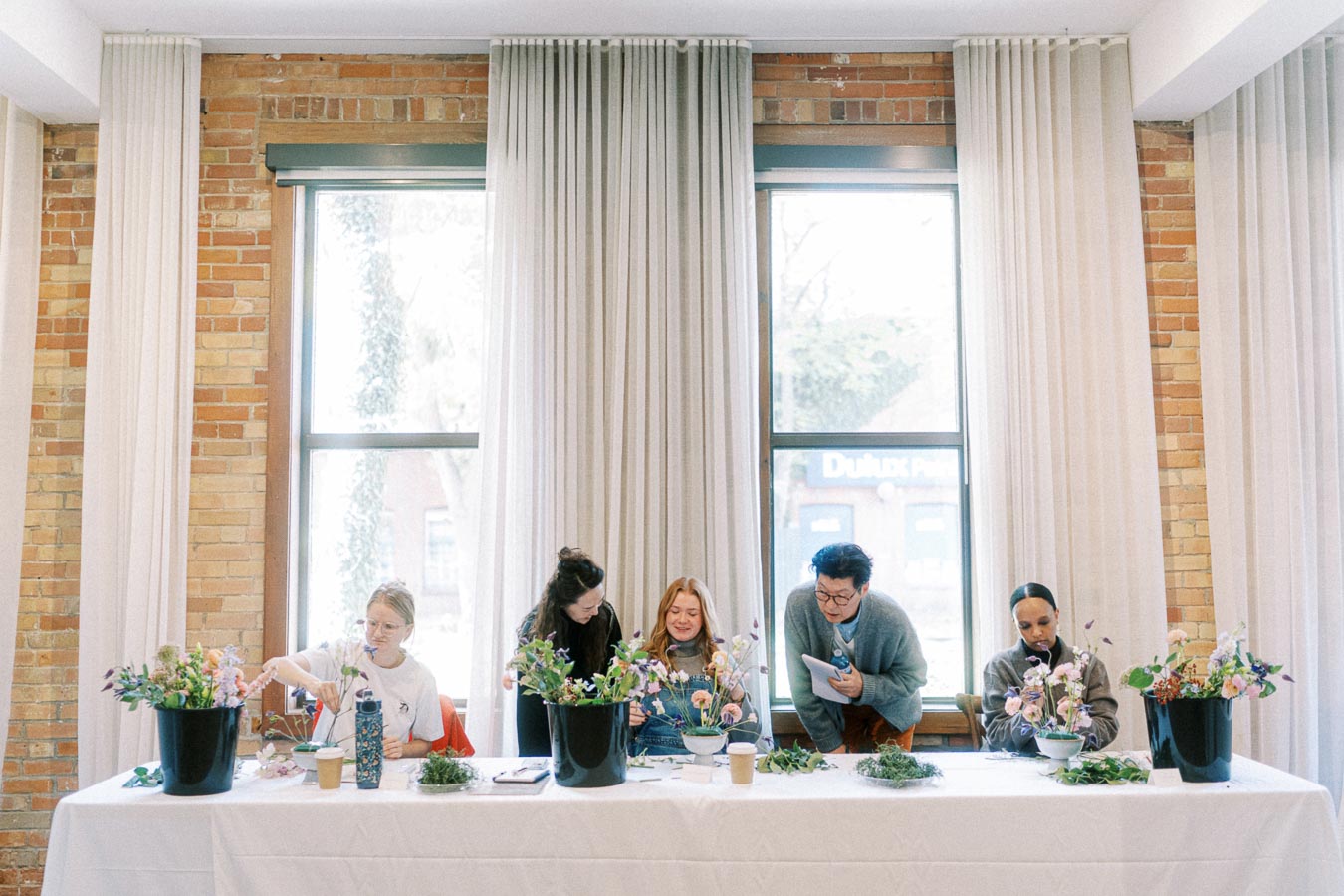 A group of people participating in a flower arrangement workshop, sitting at a table with various floral supplies, set against a backdrop of large windows and brick walls.