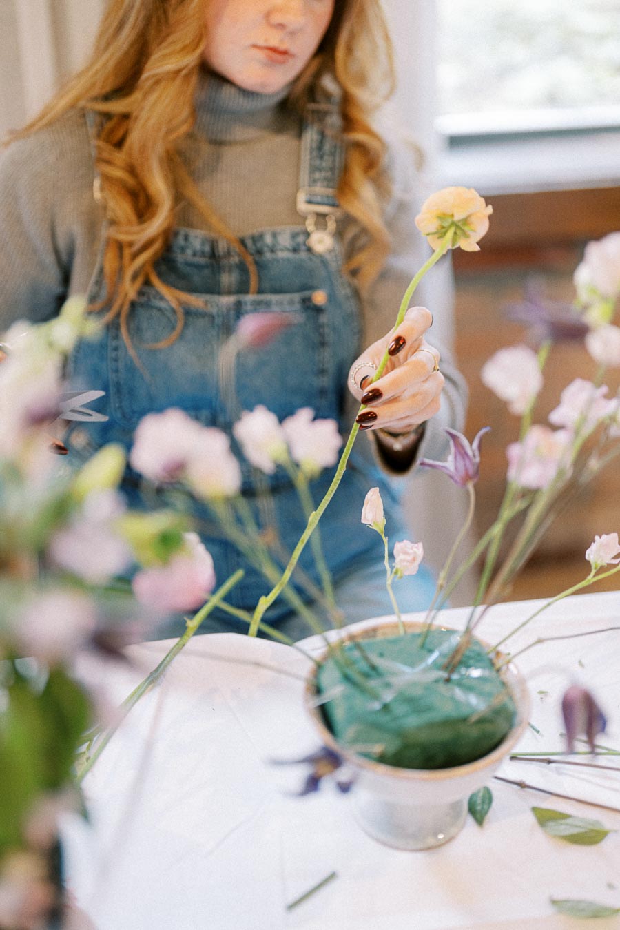 A woman arranging delicate flowers in a floral foam block, wearing a grey turtleneck and denim overalls, focused on flower arranging, soft and natural setting.