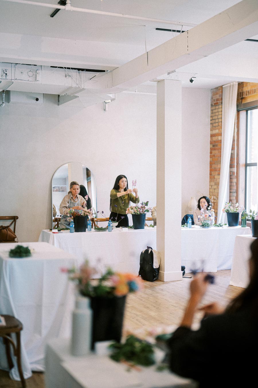 Floral arrangement workshop with participants designing bouquets in a well-lit studio space, featuring white tables covered with flower materials.