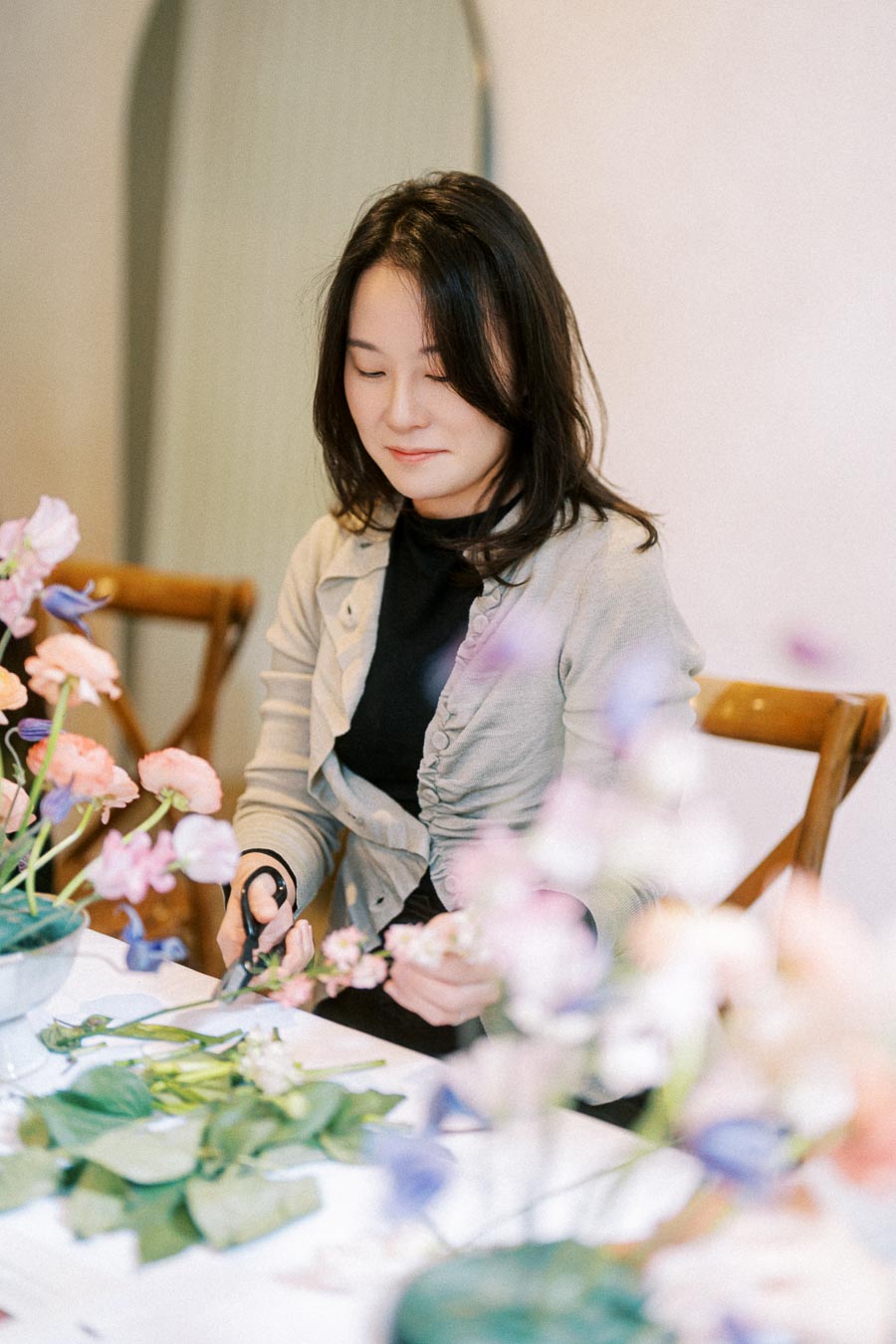 A person arranging a floral bouquet at a table, surrounded by pink and purple flowers, in a cozy indoor setting.