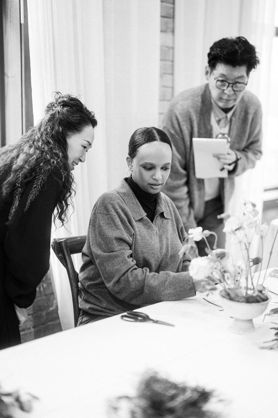 Three people collaborating in a floral design workshop, with one person seated arranging flowers, while the other two observe and suggest ideas, capturing teamwork and creativity in a bright setting.
