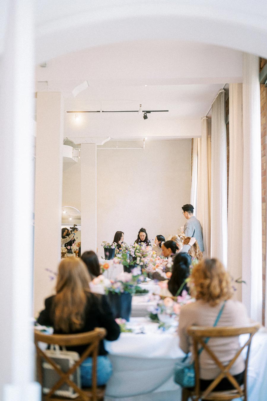 Group of people enjoying a social event in an elegantly decorated room with a long table adorned with colorful flower arrangements.