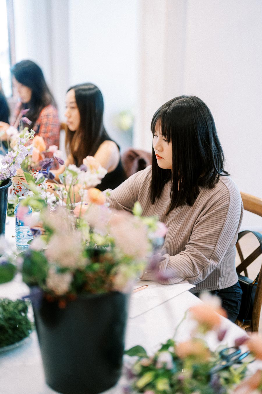 Women participating in a floral arrangement class, focusing on creating beautiful bouquets at a table filled with vibrant flowers and greenery.