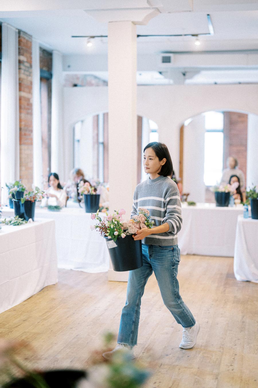 Person carrying a bucket of flowers in a floral workshop setting, surrounded by tables and natural light.