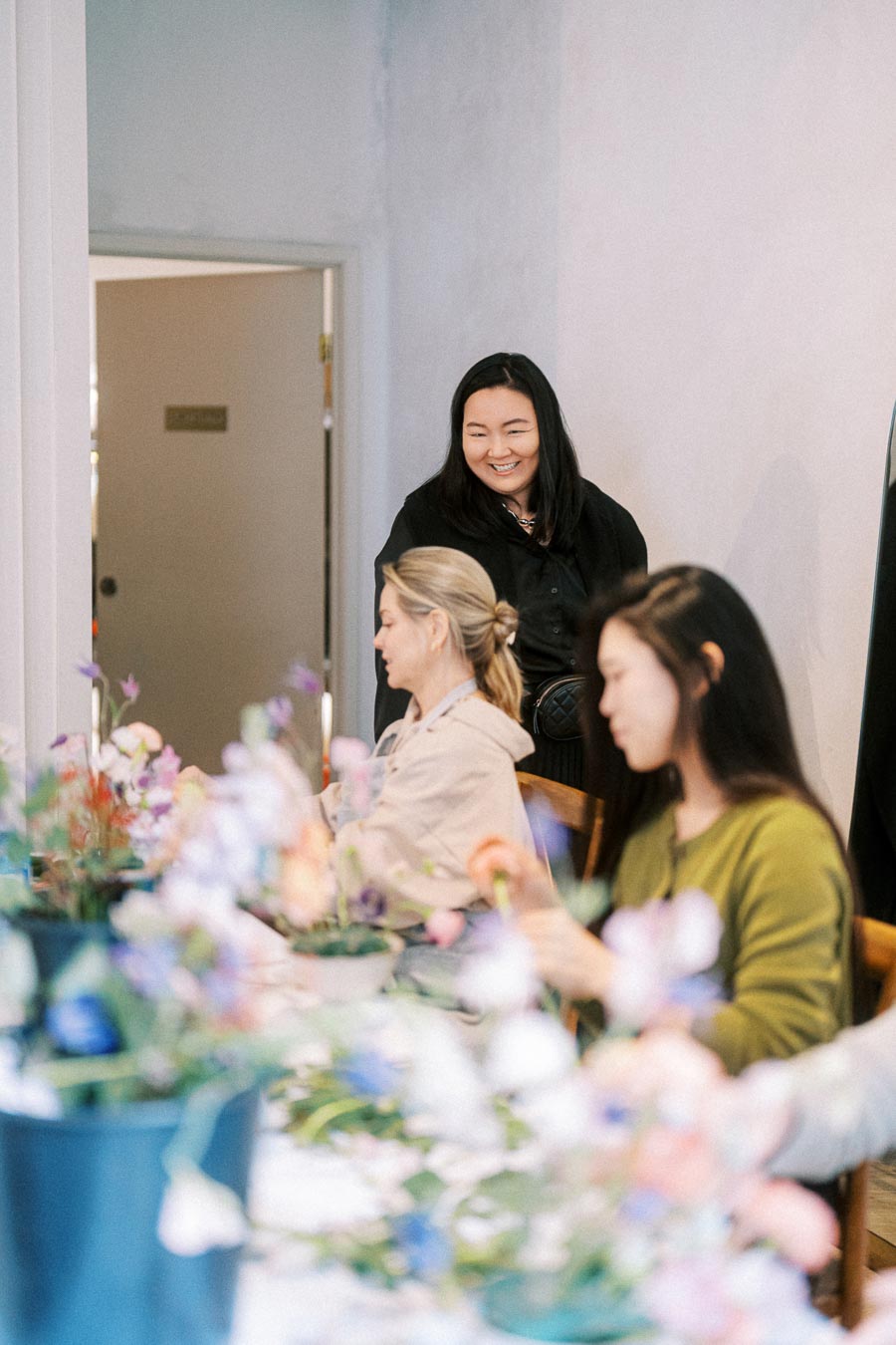 Group of women engaged in flower arranging at a workshop, with colorful blooms on the table and one woman smiling in the background.