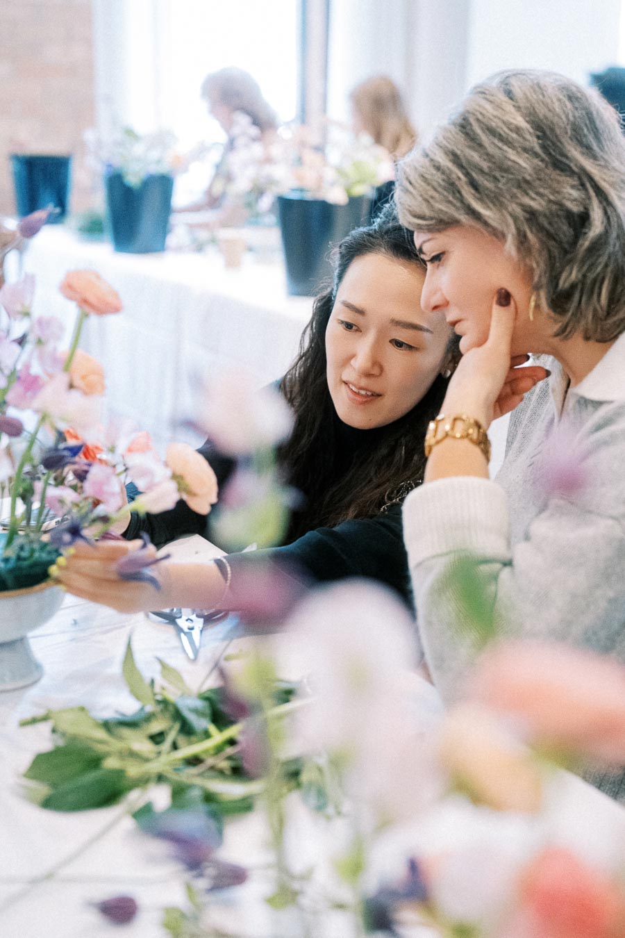 Two women engaged in a floral arrangement workshop, carefully arranging colorful flowers on a table. The setting is bright and creative, with various flowers and tools visible on the table, indicating a hands-on learning environment.
