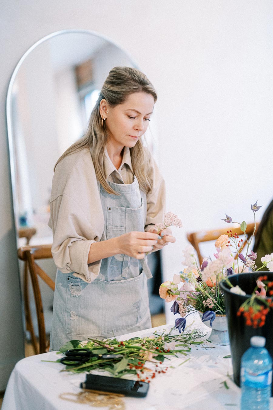 Florist arranging a colorful bouquet of flowers on a table, wearing a light apron, with a variety of floral materials and tools around her in a bright room with a mirror in the background.
