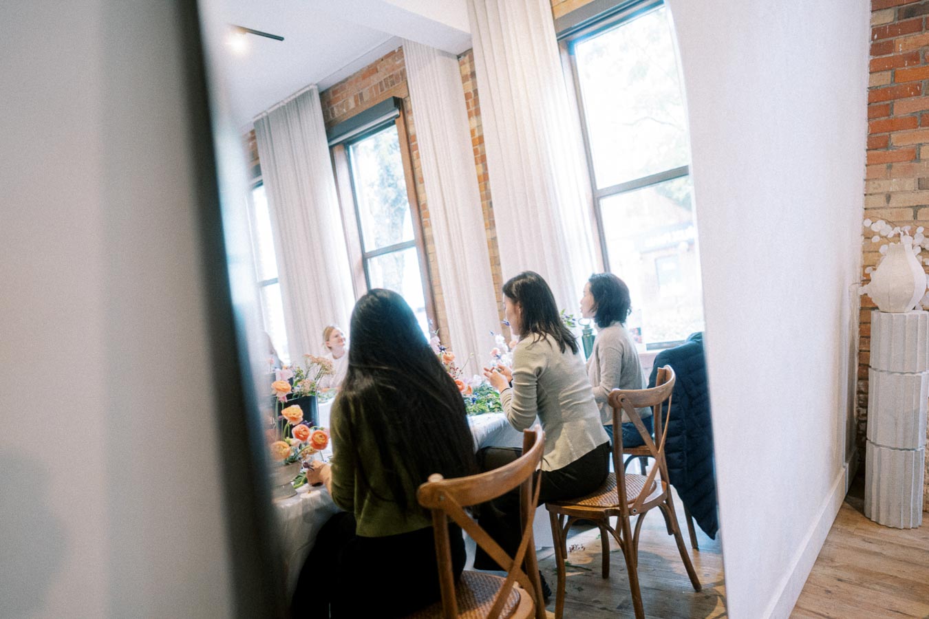 A group of women sitting at a wooden table, engaged in a meeting or workshop in a bright, modern room with large windows and brick walls, featuring floral decorations.