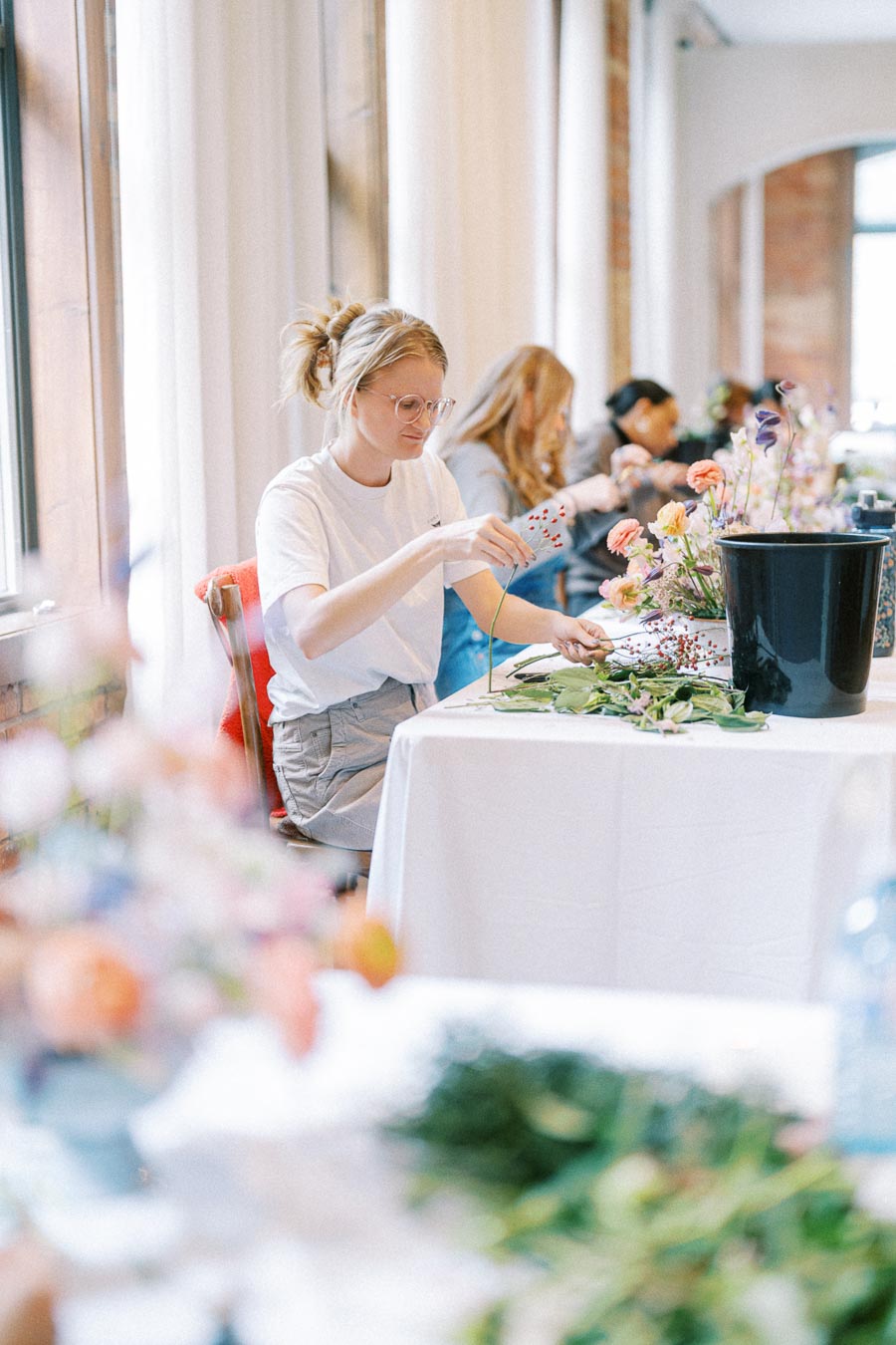 A woman in a white t-shirt and glasses arranges flowers on a table during a floral decoration workshop, with various colorful blooms and leaves.
