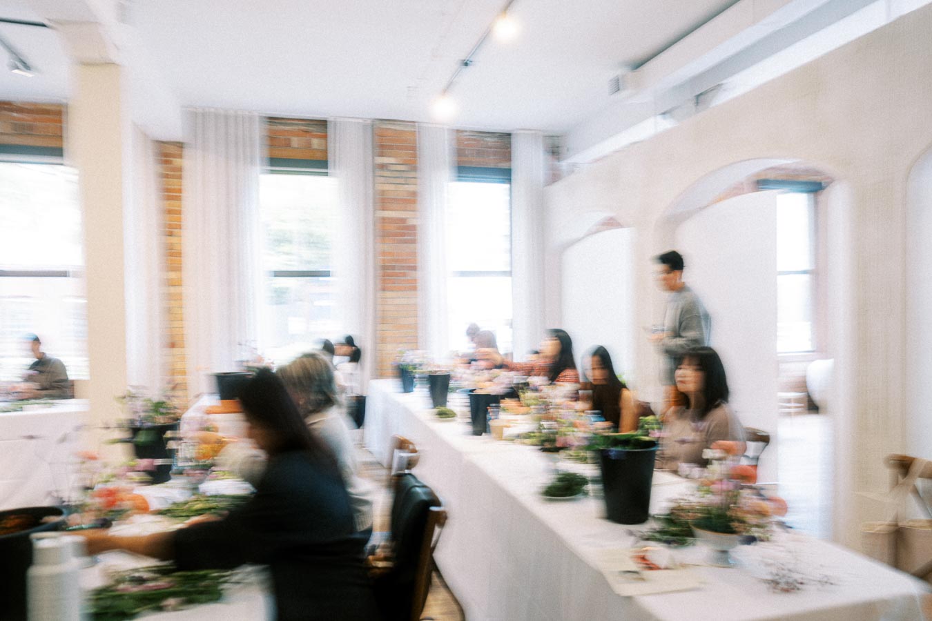 A group of people seated at long tables in a bright, modern room with brick accents, engaged in a workshop surrounded by flowers and plants.