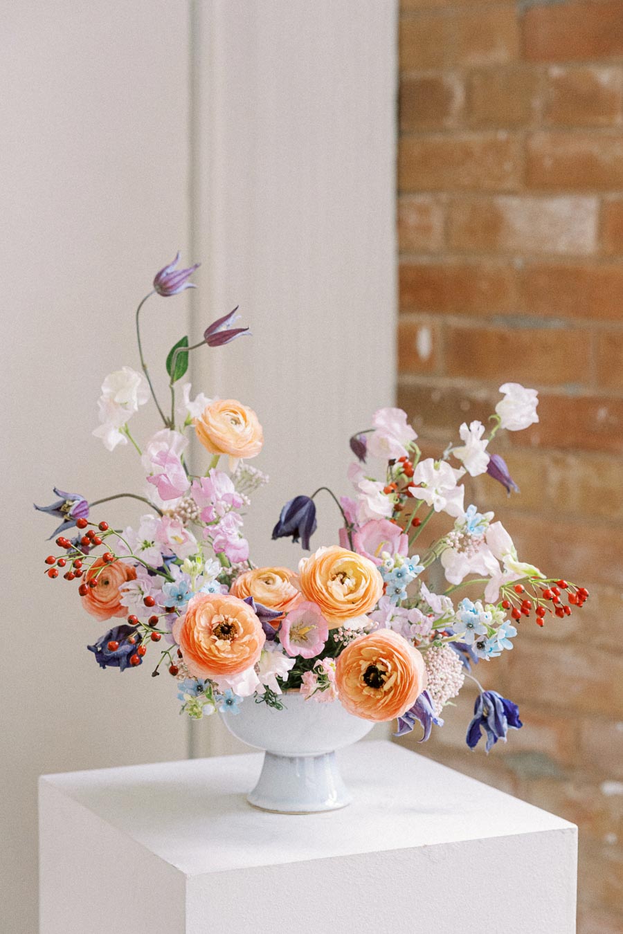 Elegant floral arrangement featuring a variety of colorful flowers, including orange ranunculus, pink and white blossoms, and purple accents in a white ceramic vase, set against a neutral indoor backdrop.