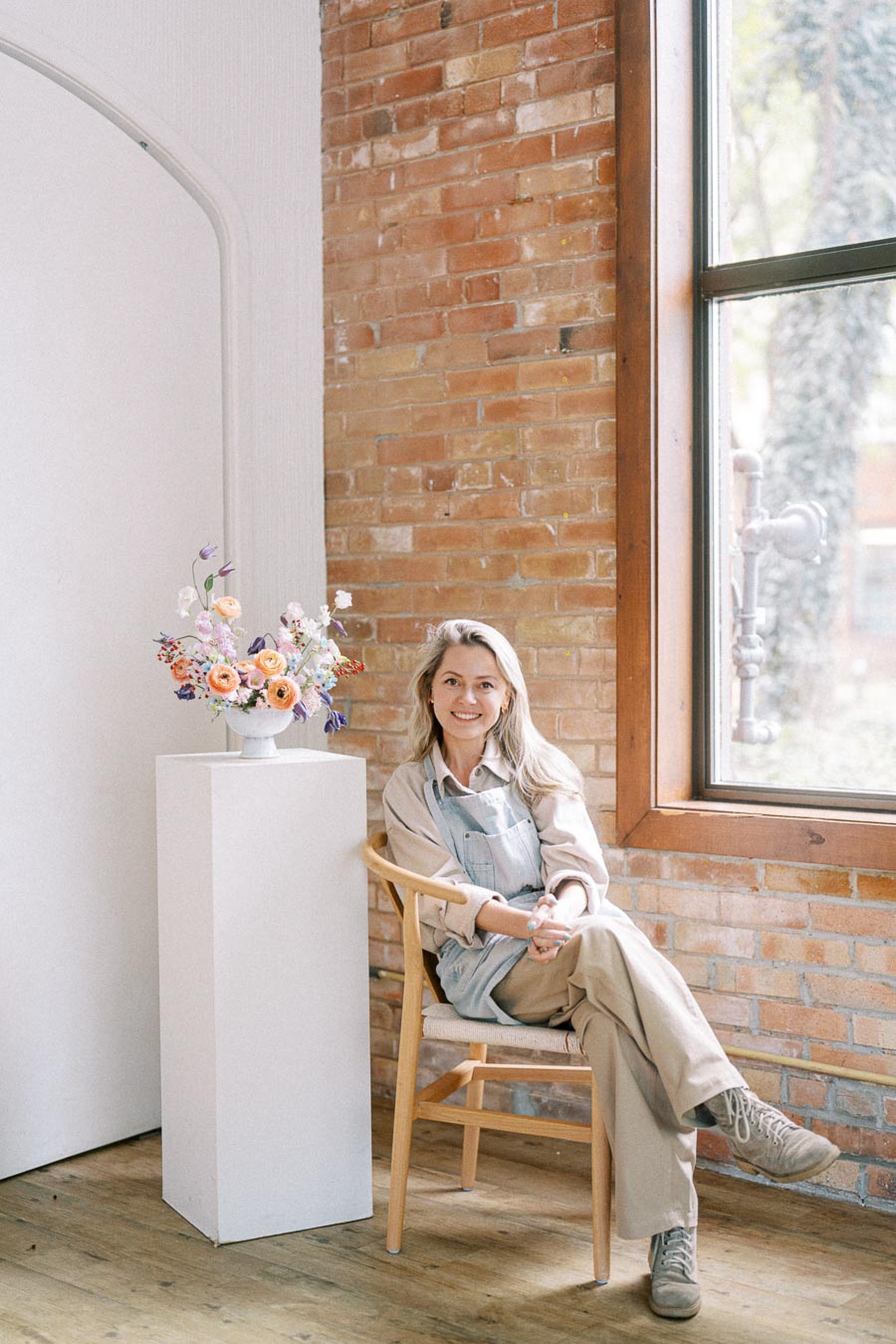 A person sitting on a wooden chair in a bright, cozy room with a brick wall and large window. They are wearing casual clothing and an apron. A white pedestal next to them displays a colorful floral arrangement.