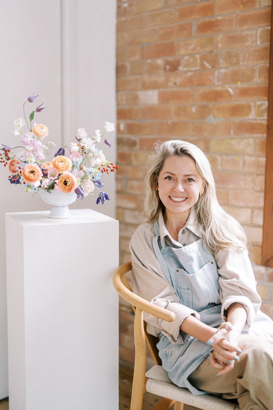 Smiling woman in an apron seated next to a decorative floral arrangement on a pedestal, with a rustic brick wall in the background.