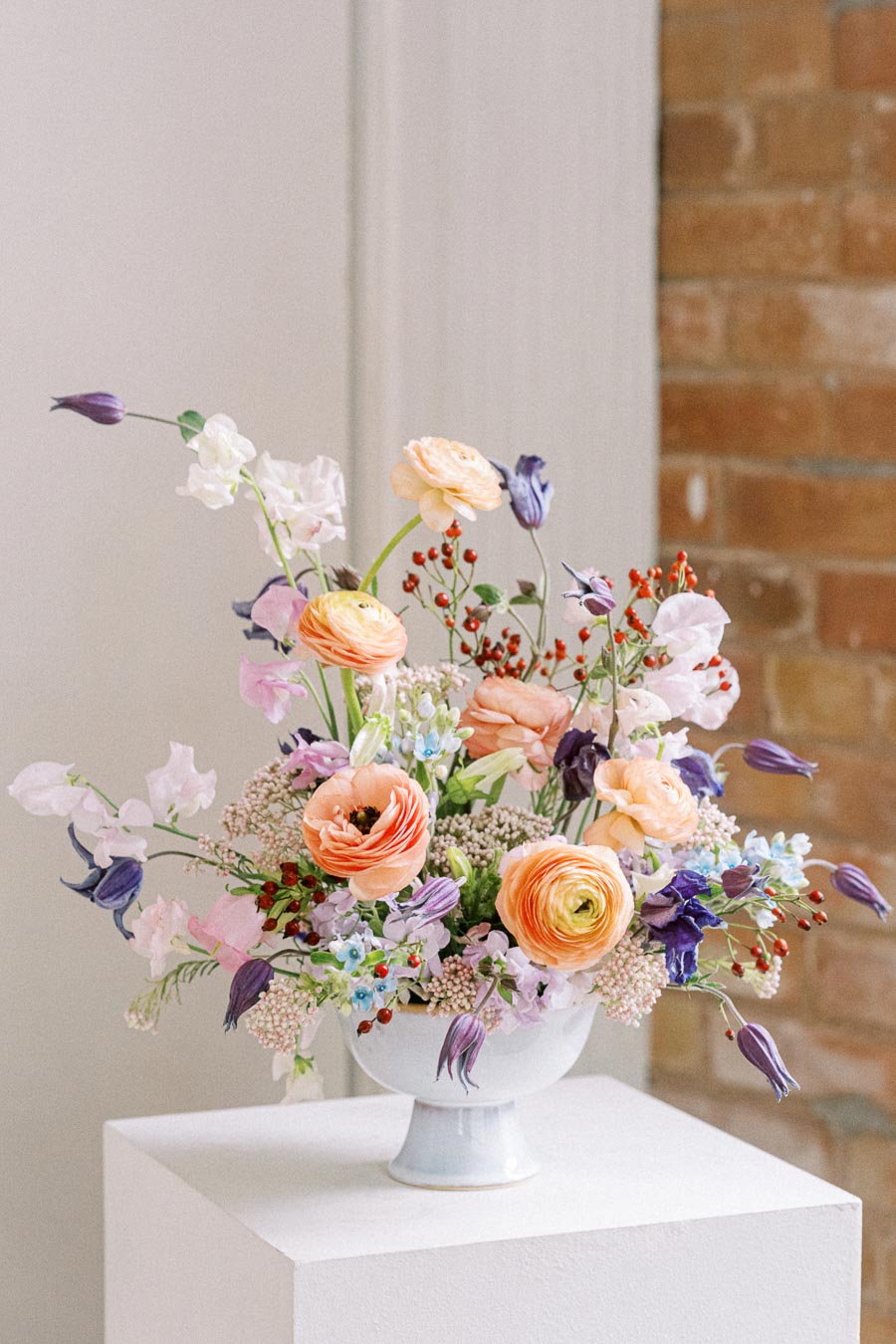 A vibrant floral arrangement in a white vase features a mix of colorful flowers including peach ranunculus, purple bellflowers, and delicate white blooms, displayed on a pedestal against a rustic brick wall.