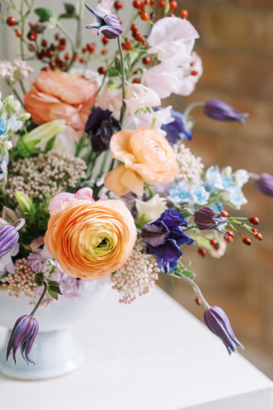 Elegant floral arrangement with peach ranunculus, purple clematis, blue delphiniums, and red berries in a white vase on a soft background.