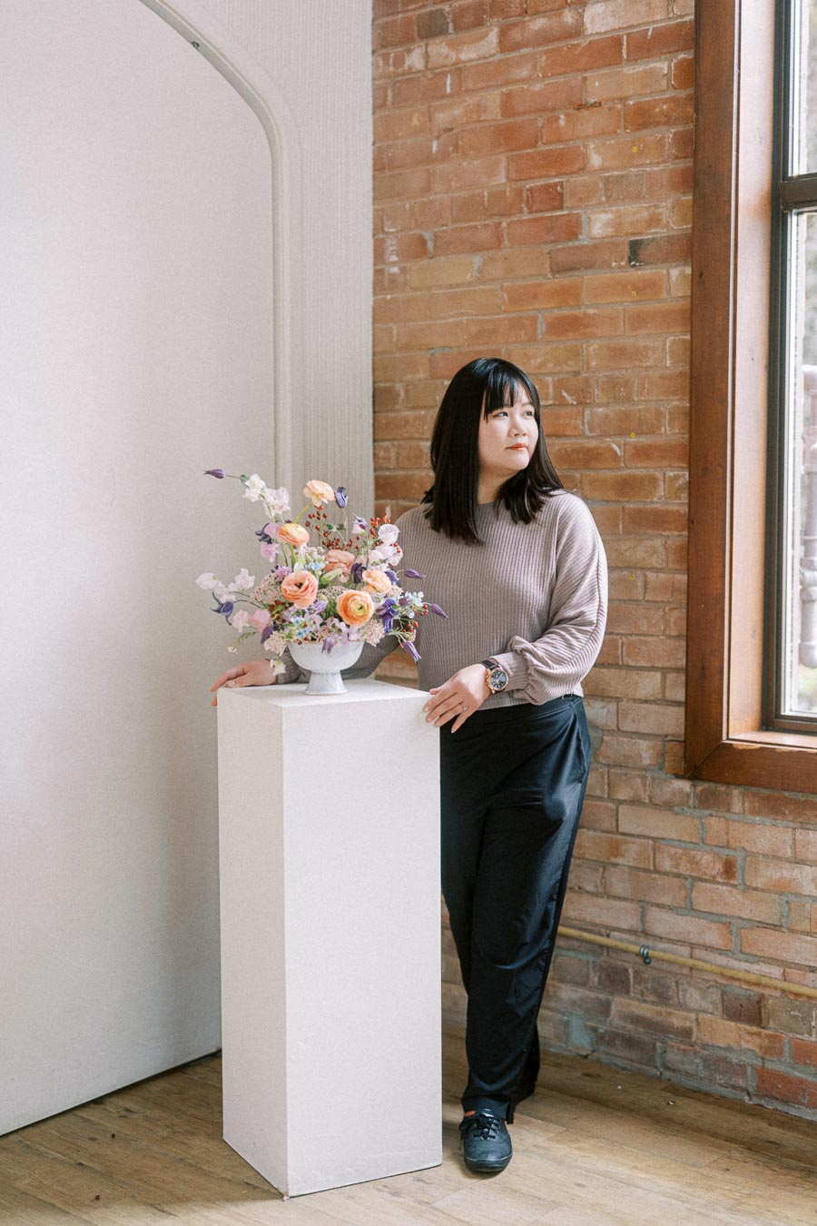 Person standing next to a floral arrangement on a pedestal in a well-lit room with brick wall and window backdrop, showcasing a blend of modern and rustic interior design.