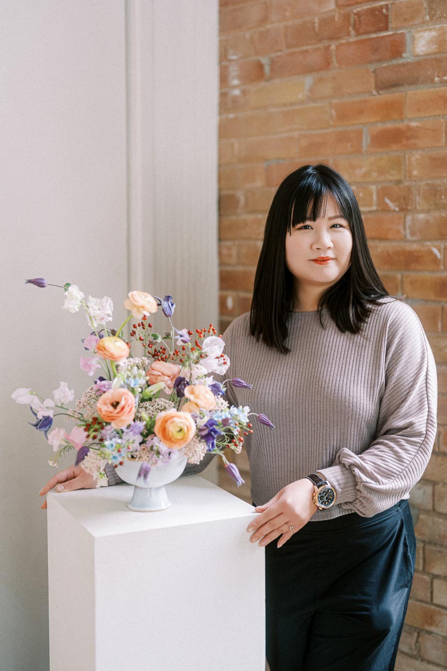 Person standing beside a pedestal with a colorful floral arrangement in a modern setting, wearing a gray sweater and black pants, showcasing flower decoration skills.