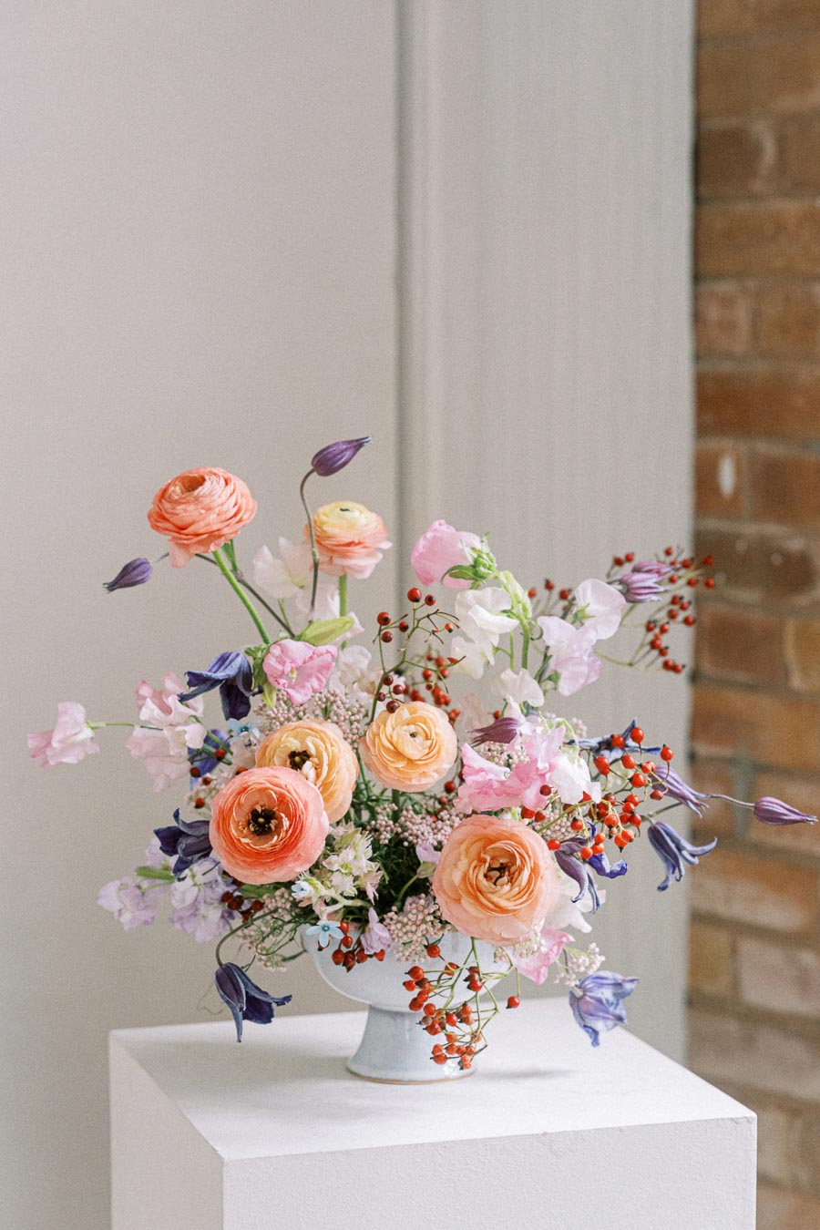 Elegant floral arrangement featuring pastel-colored flowers including pink, peach, and purple blooms with small red berries, displayed in a white vase against a soft background. Perfect for wedding or home decor inspiration.
