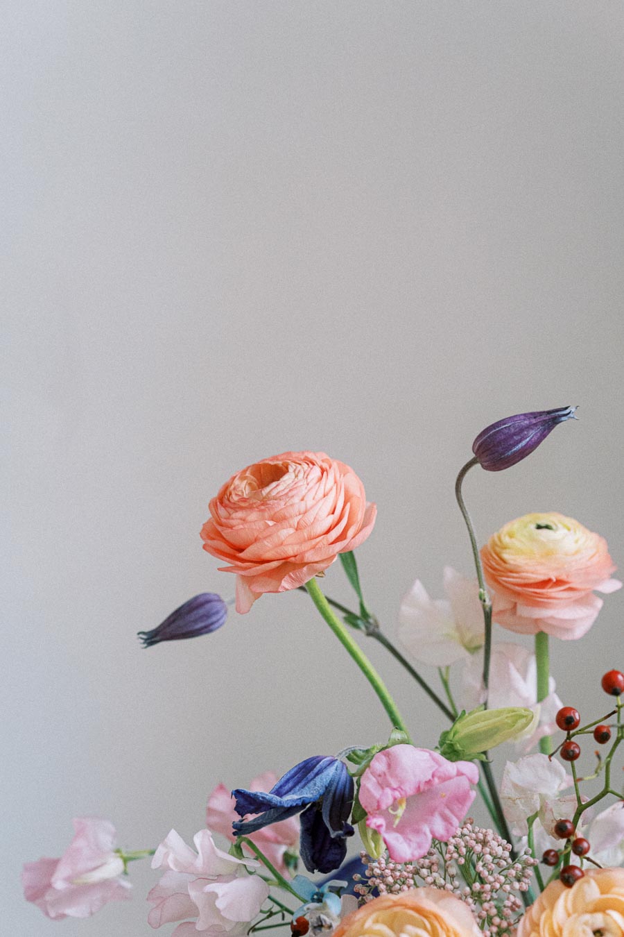 A vibrant floral arrangement featuring orange ranunculus, purple buds, and delicate pink and white blossoms against a soft gray background. Perfect for spring decoration or wedding inspiration.
